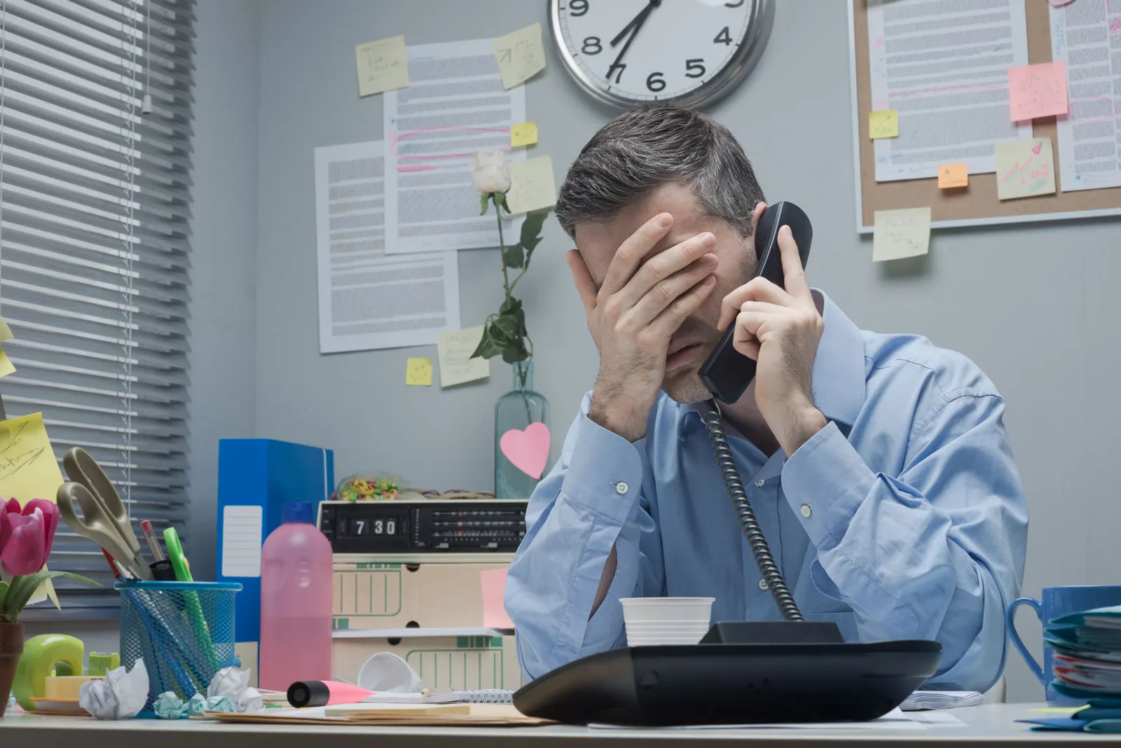A busy male office worker is stressfully holding his head in one hand and holding a phone in the other.