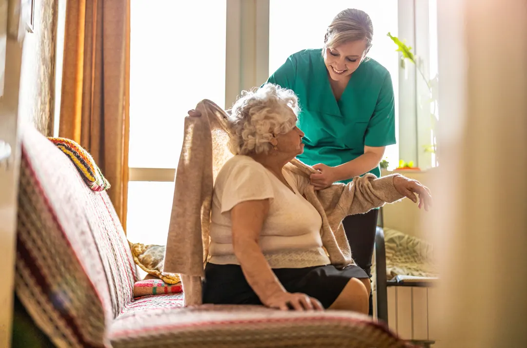 A female nurse helping an elderly patient put on a sweater inside of her home.