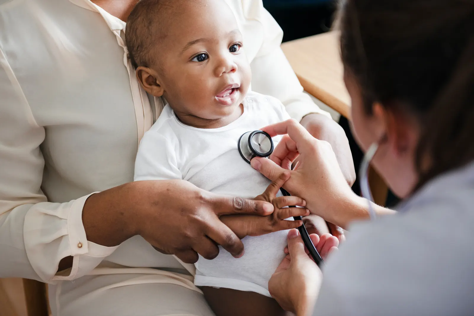 A doctor checking the heartbeat of a baby. The baby is being held in their mothers arms while the doctor closely listens in with her stethoscope.