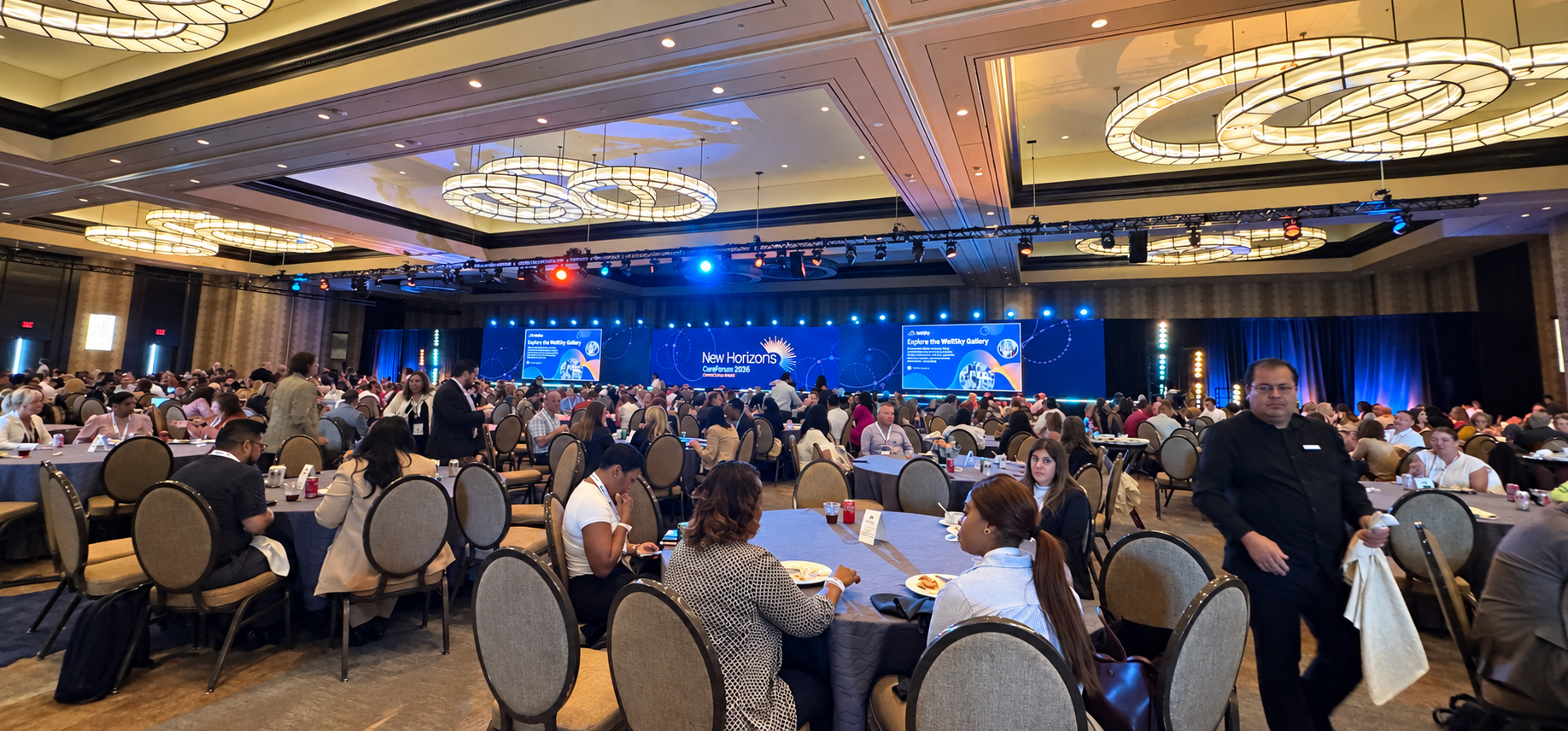 Attendees seated at round tables in the main ballroom at WellSky CareForum 2026, with the New Horizons conference branding displayed on stage screens